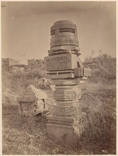 A single thick stone pillar, carved in bands with a broken top, standing in rough grass among fallen carved blocks, with low ruins behind.