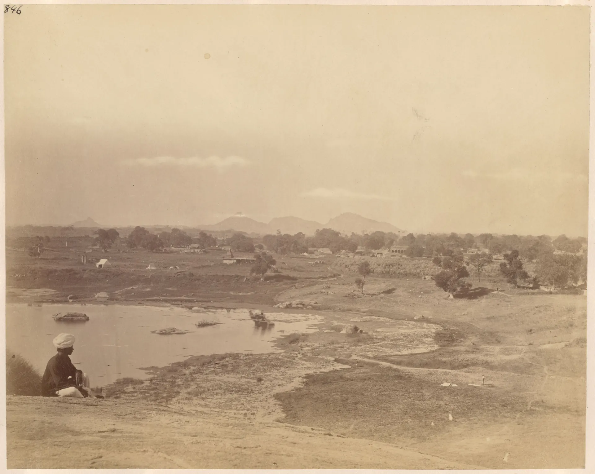 A wide view from a rocky height over a stretch of water and open fields with scattered trees and huts, low hills on the horizon and a man seated in the left foreground.