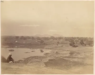 A wide view from a rocky height over a stretch of water and open fields with scattered trees and huts, low hills on the horizon and a man seated in the left foreground.