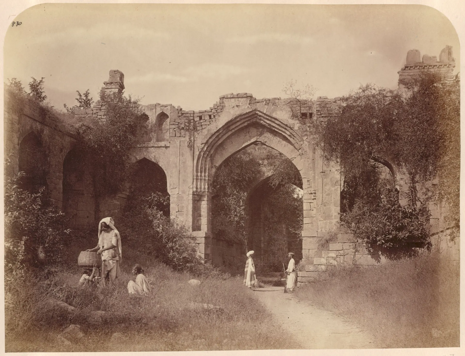 A ruined gateway with a tall pointed arch and side arches, overgrown with plants, with a path running through it and people standing in front.