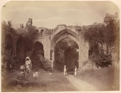 A ruined gateway with a tall pointed arch and side arches, overgrown with plants, with a path running through it and people standing in front.