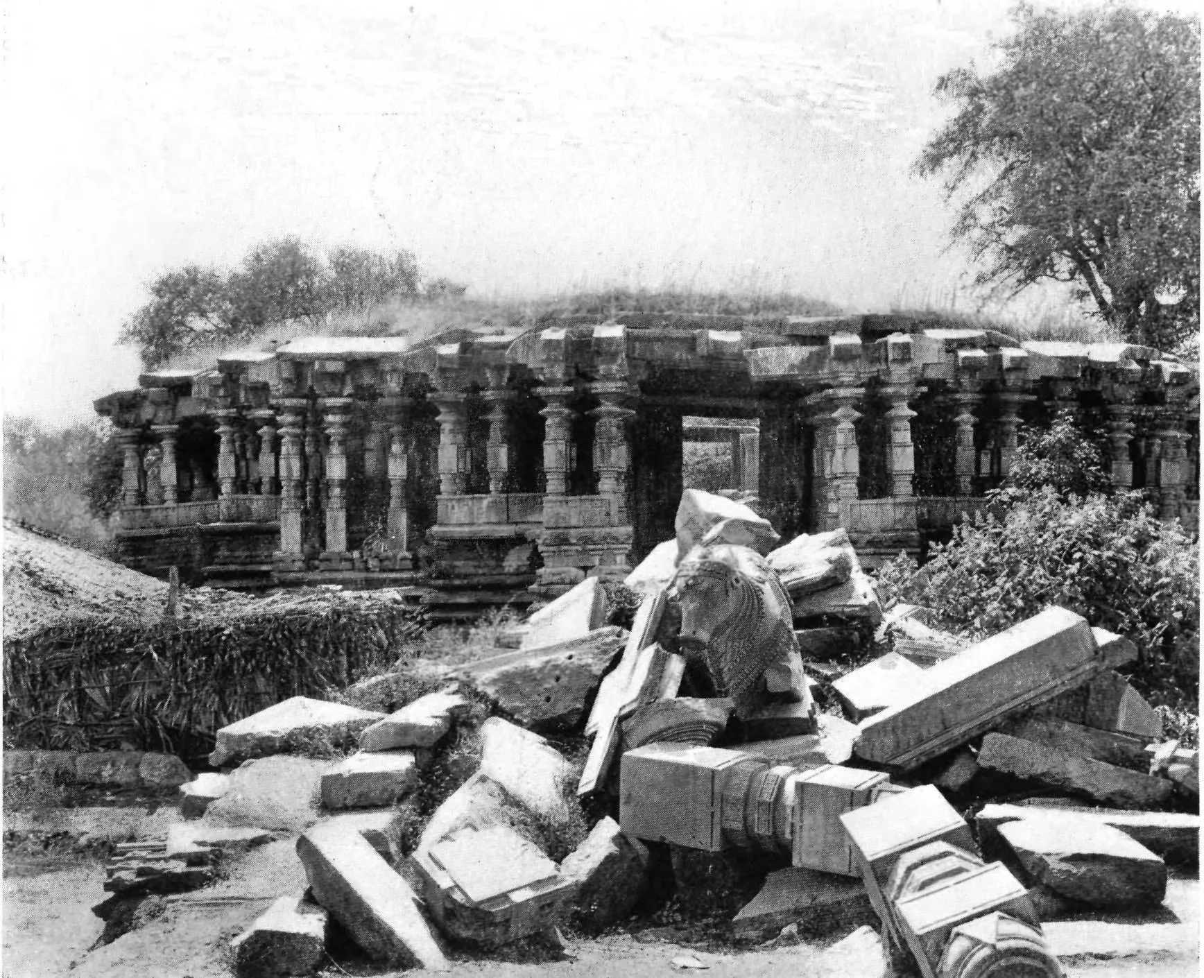 Photograph of a long, low pillared stone hall with a flat roof, a jumble of fallen carved blocks and pillars heaped in the foreground and trees at the right.