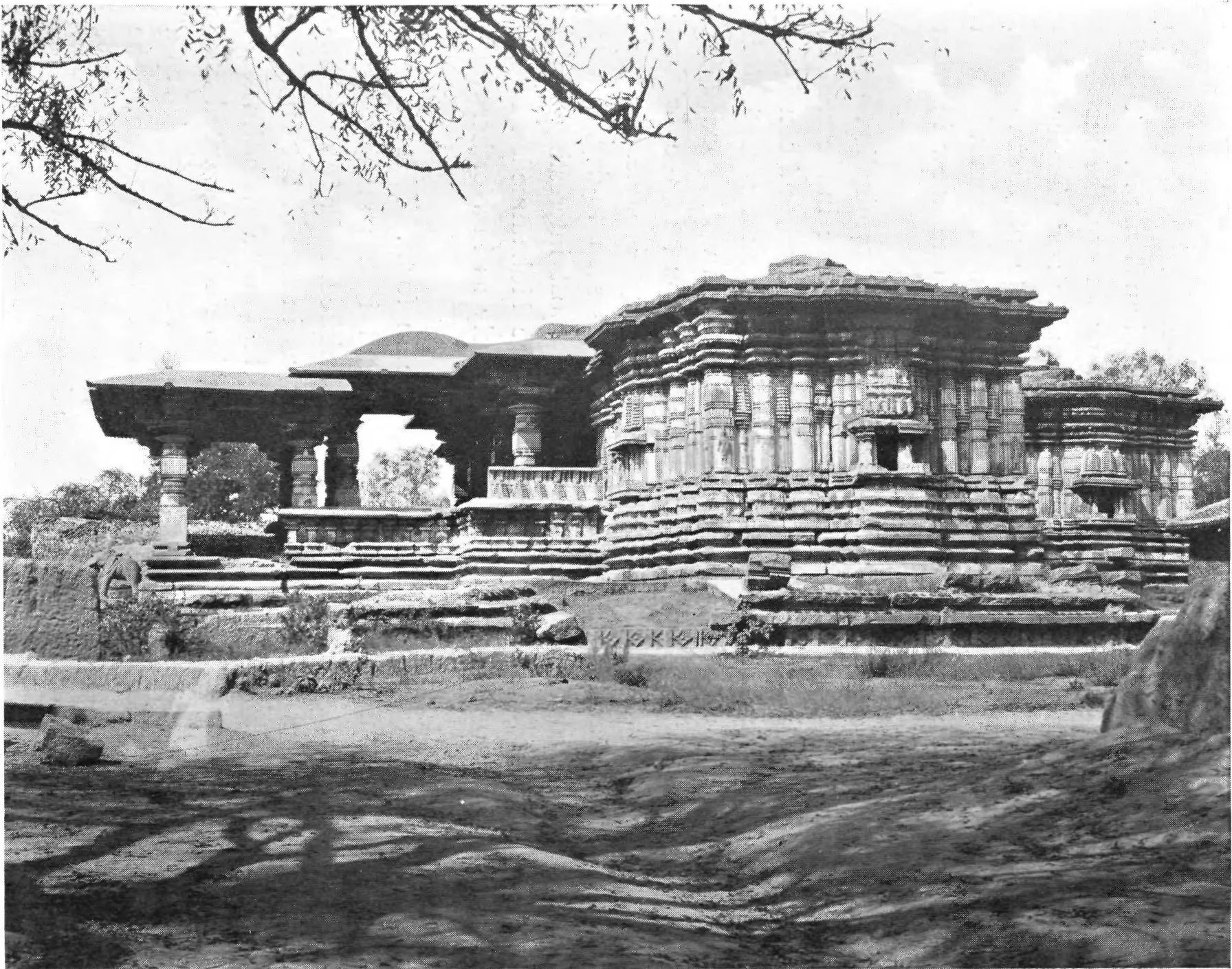 Photograph of a low, wide stone temple on a moulded platform with flat-roofed pillared porches and stepped walls, seen across open ground, with tree branches above.