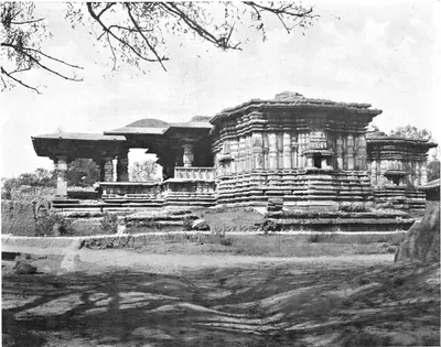 Photograph of a low, wide stone temple on a moulded platform with flat-roofed pillared porches and stepped walls, seen across open ground, with tree branches above.