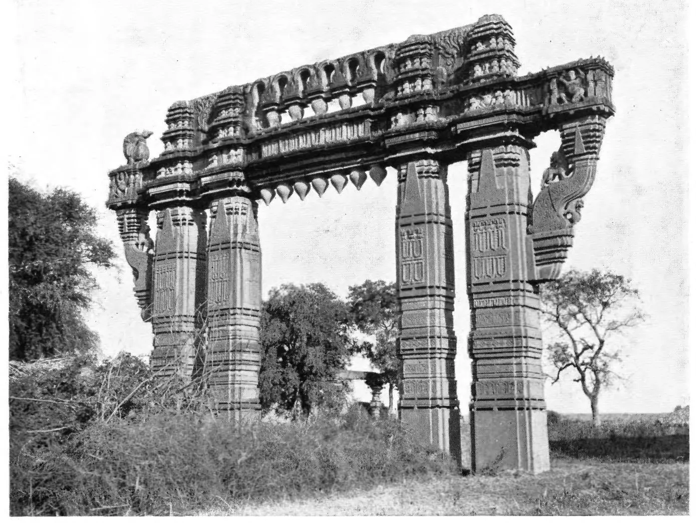 Photograph of a tall stone gateway of carved pillars and a deep sculptured lintel standing among bushes and trees, seen at an angle.