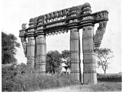 Photograph of a tall stone gateway of carved pillars and a deep sculptured lintel standing among bushes and trees, seen at an angle.