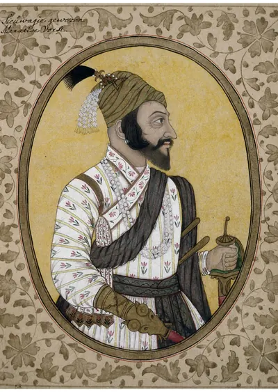 Deccani bust-length portrait of a bearded man in profile with a plumed turban, a striped robe and a dark sash, holding a sword hilt, in an oval frame on a yellow ground within a floral border.