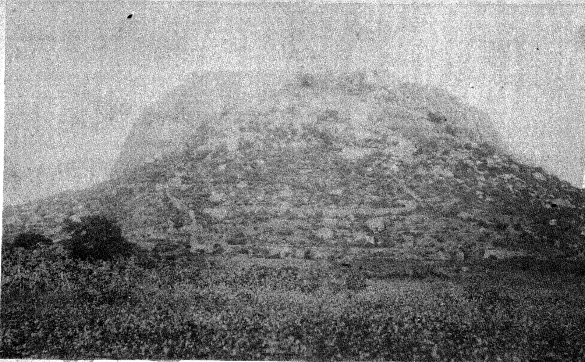 Faded black-and-white photograph of a large rounded rocky hill rising above scrub-covered ground, with faint lines of walls across its slopes.