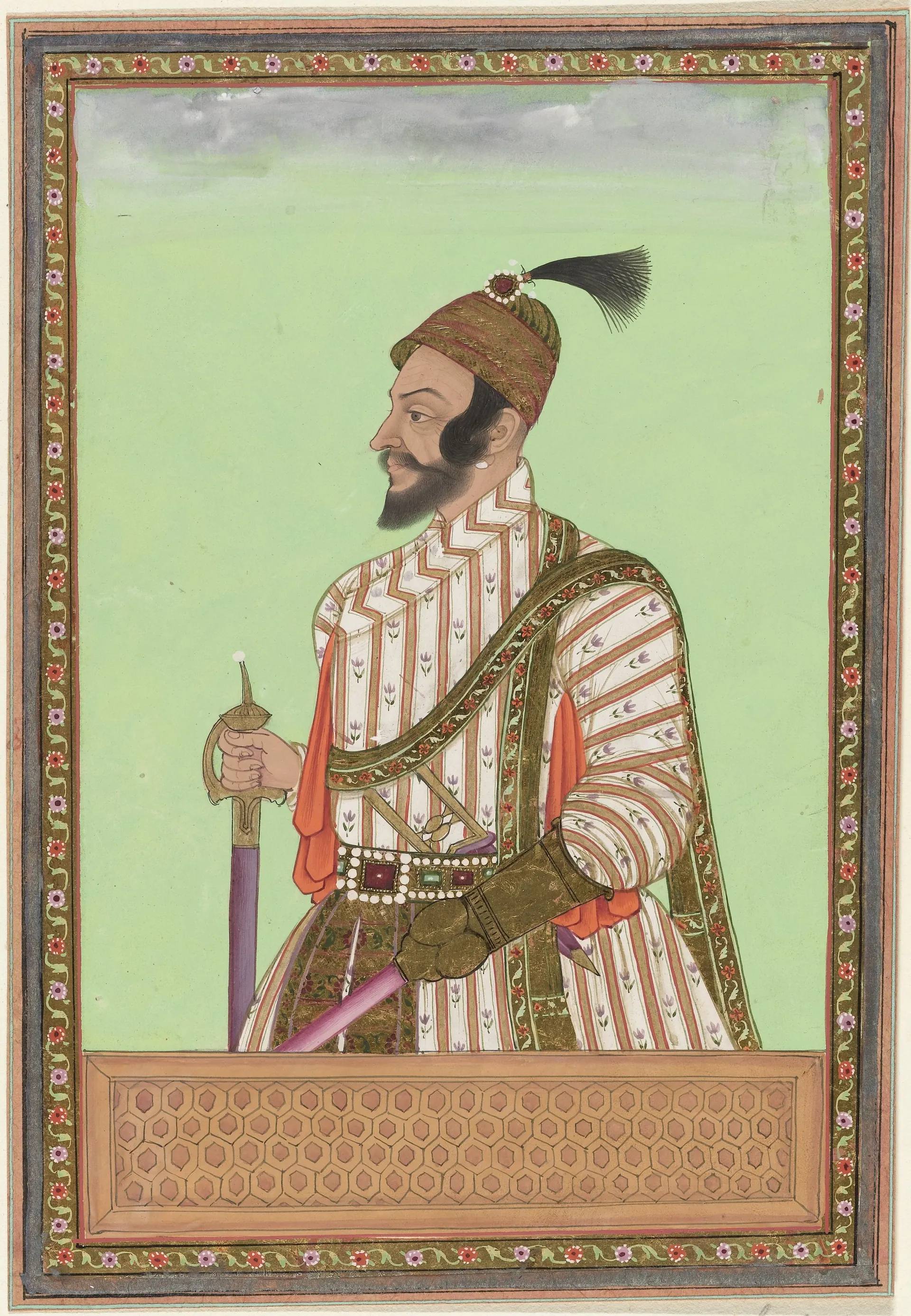 Deccani painting of a bearded man in a striped robe and plumed turban, a sword in his right hand and a lance in his left, standing behind a decorated parapet against a green ground, in a floral border.