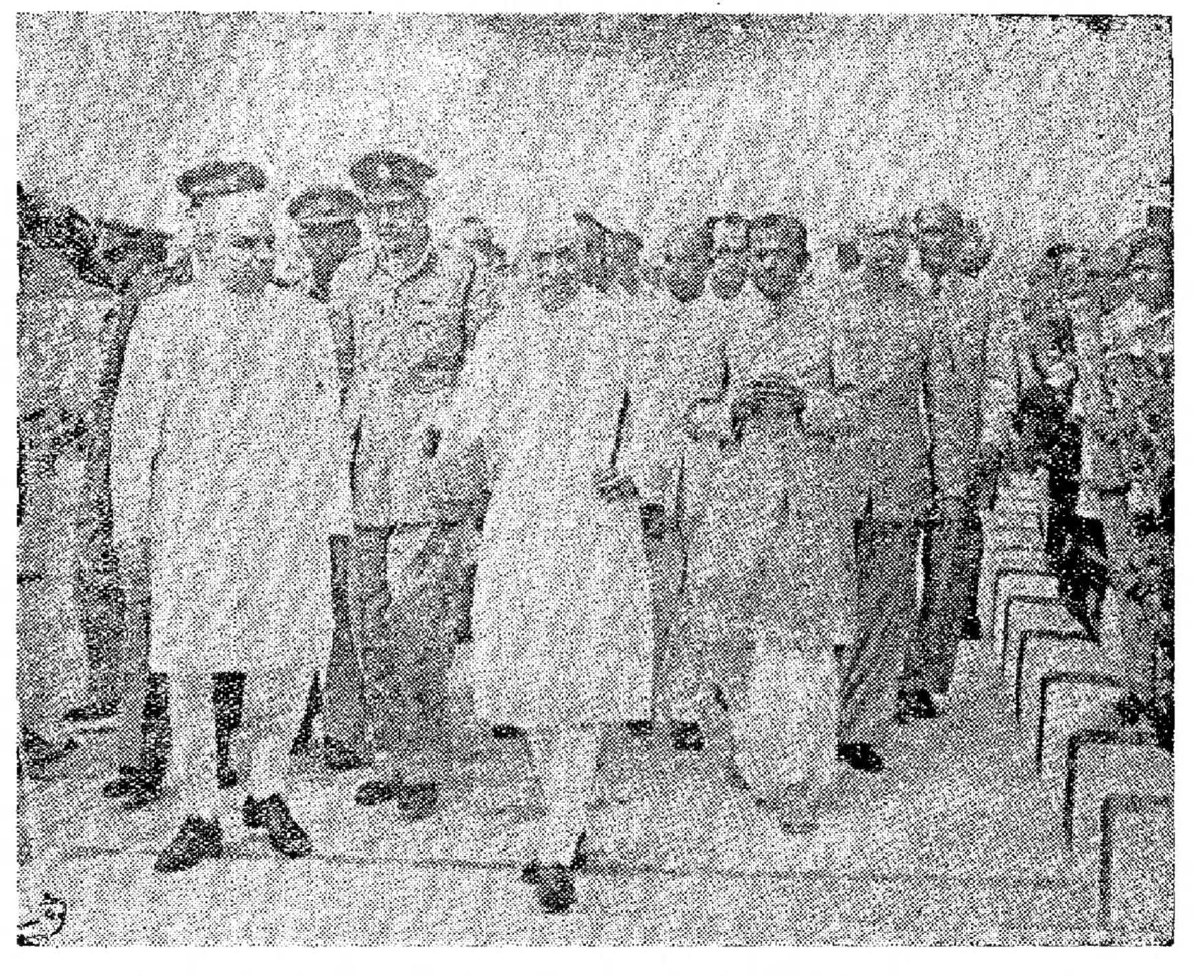 A grainy black-and-white photograph of a group of men in white clothes and caps walking towards the camera between rows of people, with uniformed officers among them.