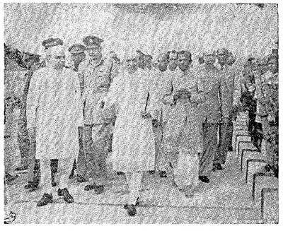 A grainy black-and-white photograph of a group of men in white clothes and caps walking towards the camera between rows of people, with uniformed officers among them.