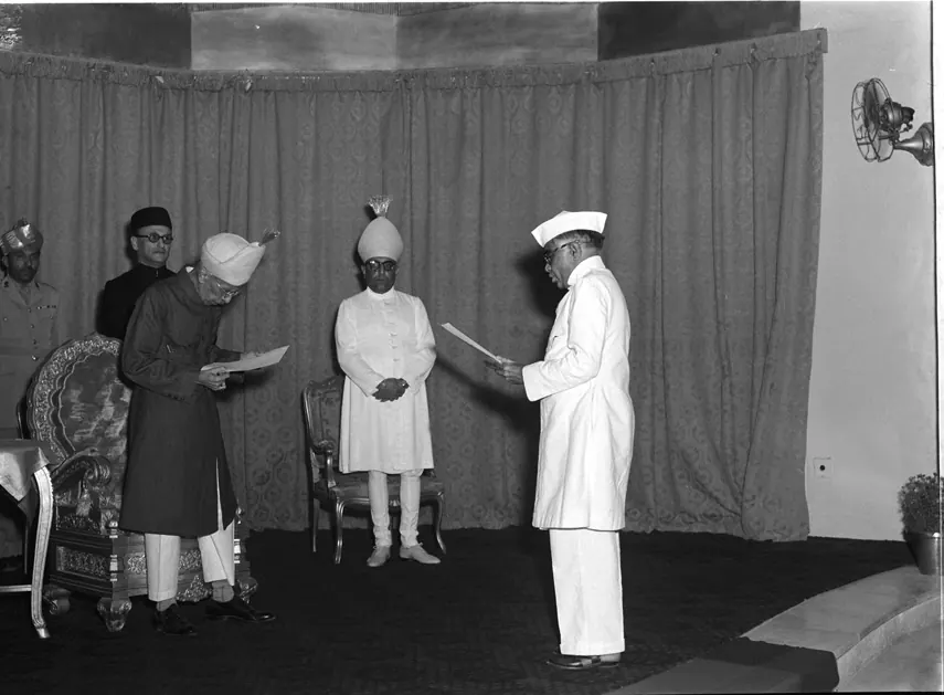 A black-and-white photograph of a man in a white cap and coat reading from a paper before an older man in a dark long coat and cap who holds a paper, in a curtained room with a few others standing behind.