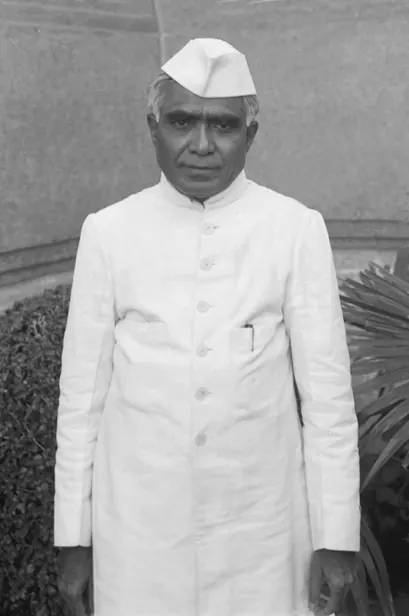 A black-and-white photograph of a man in a white cap and a long white buttoned coat, standing in front of a wall and a bush.