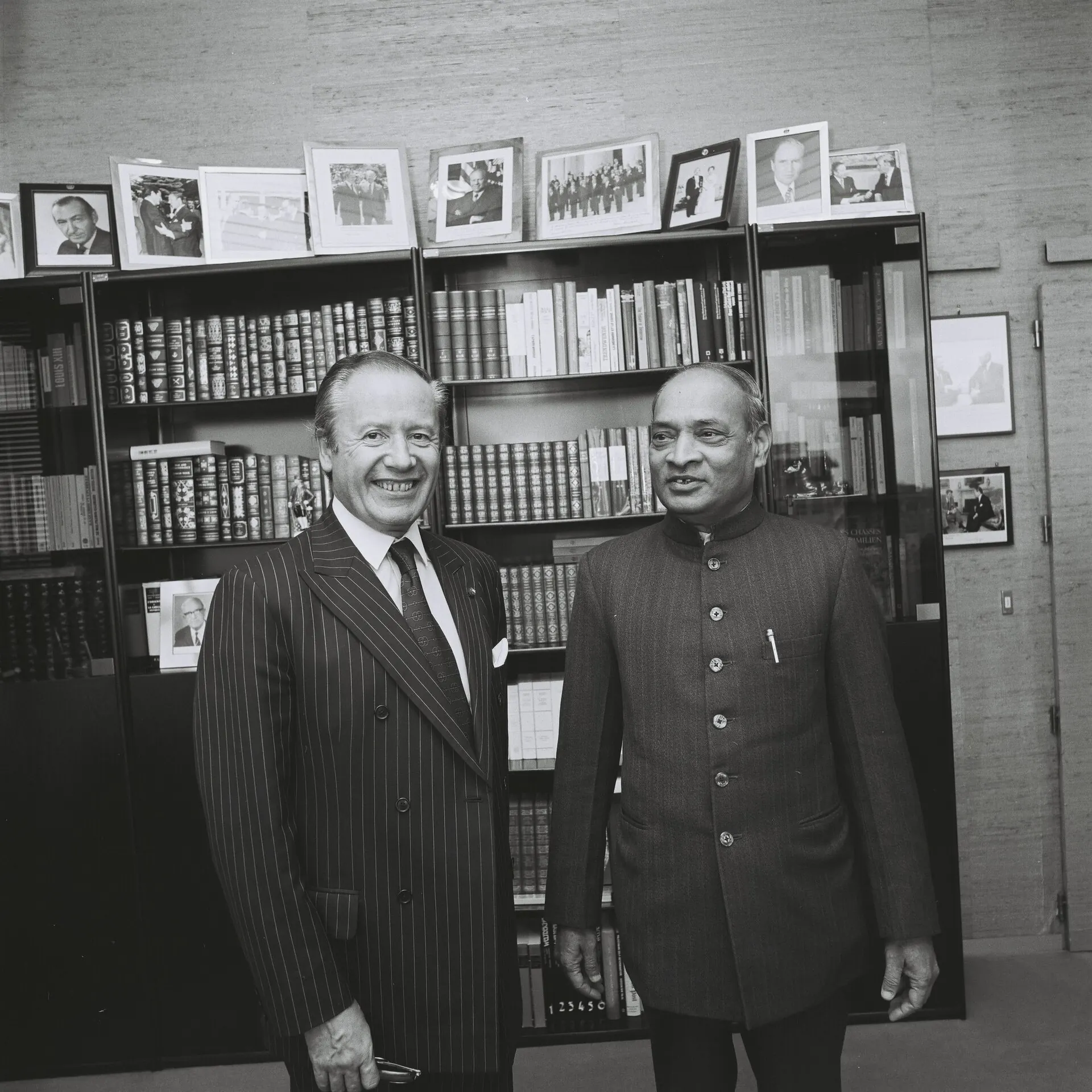 A black-and-white photograph of two men standing and smiling in an office in front of glass-fronted bookcases with framed photographs on top; the man on the right wears a long buttoned Indian jacket, the man on the left a pinstriped suit.