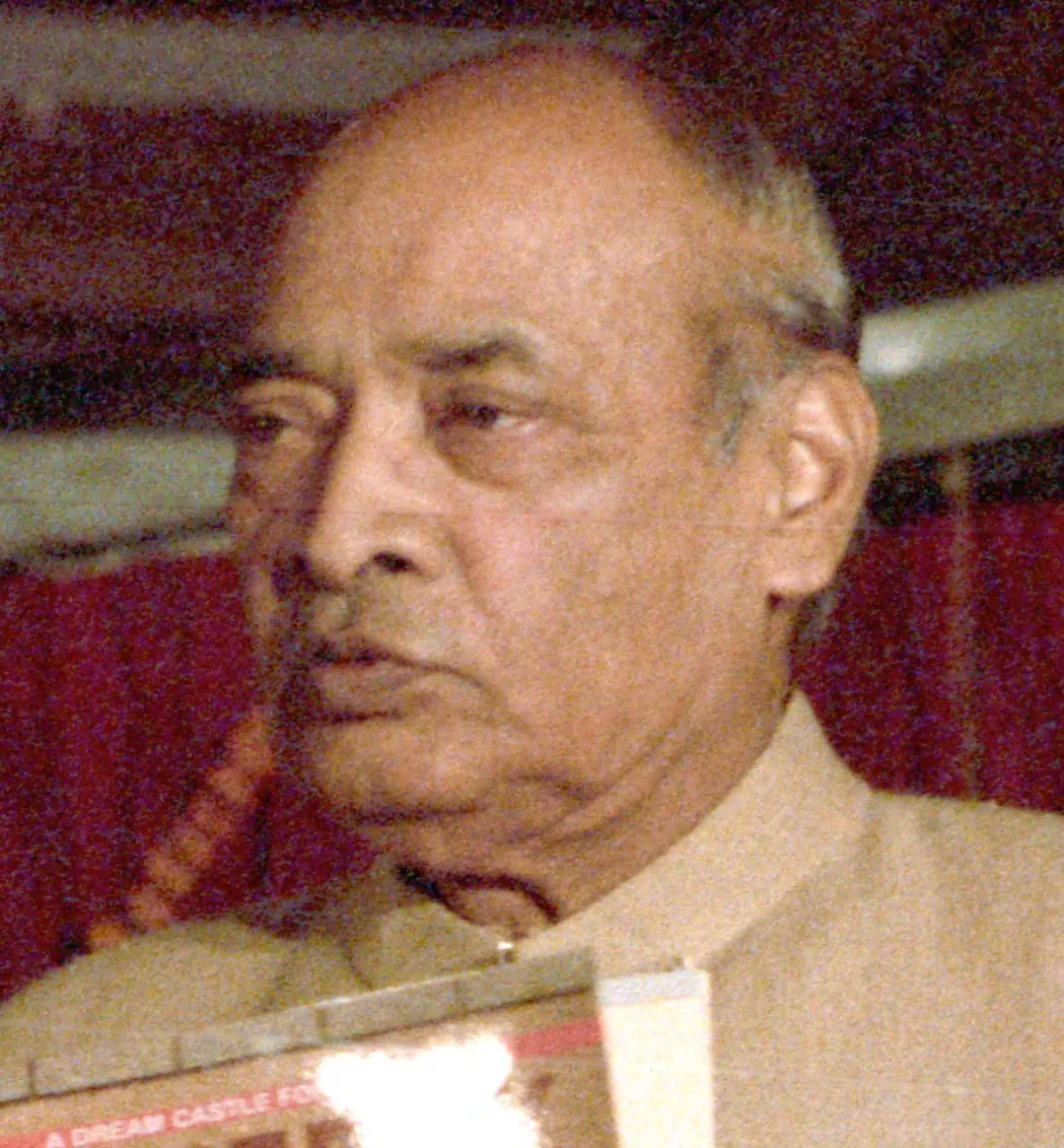 A colour close-up of an elderly bald man with a heavy lower lip, in a cream-coloured jacket, looking to the left, with a red background.