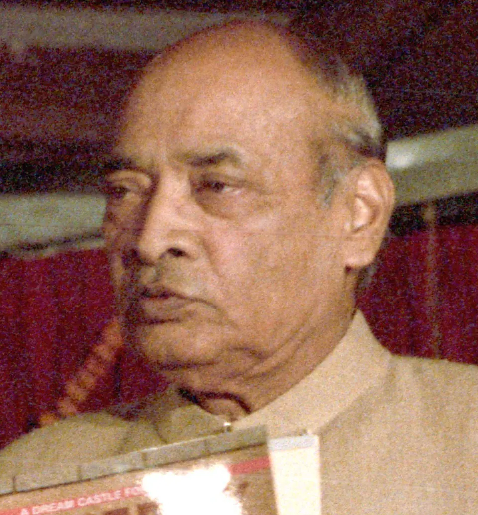 A colour close-up of an elderly bald man with a heavy lower lip, in a cream-coloured jacket, looking to the left, with a red background.