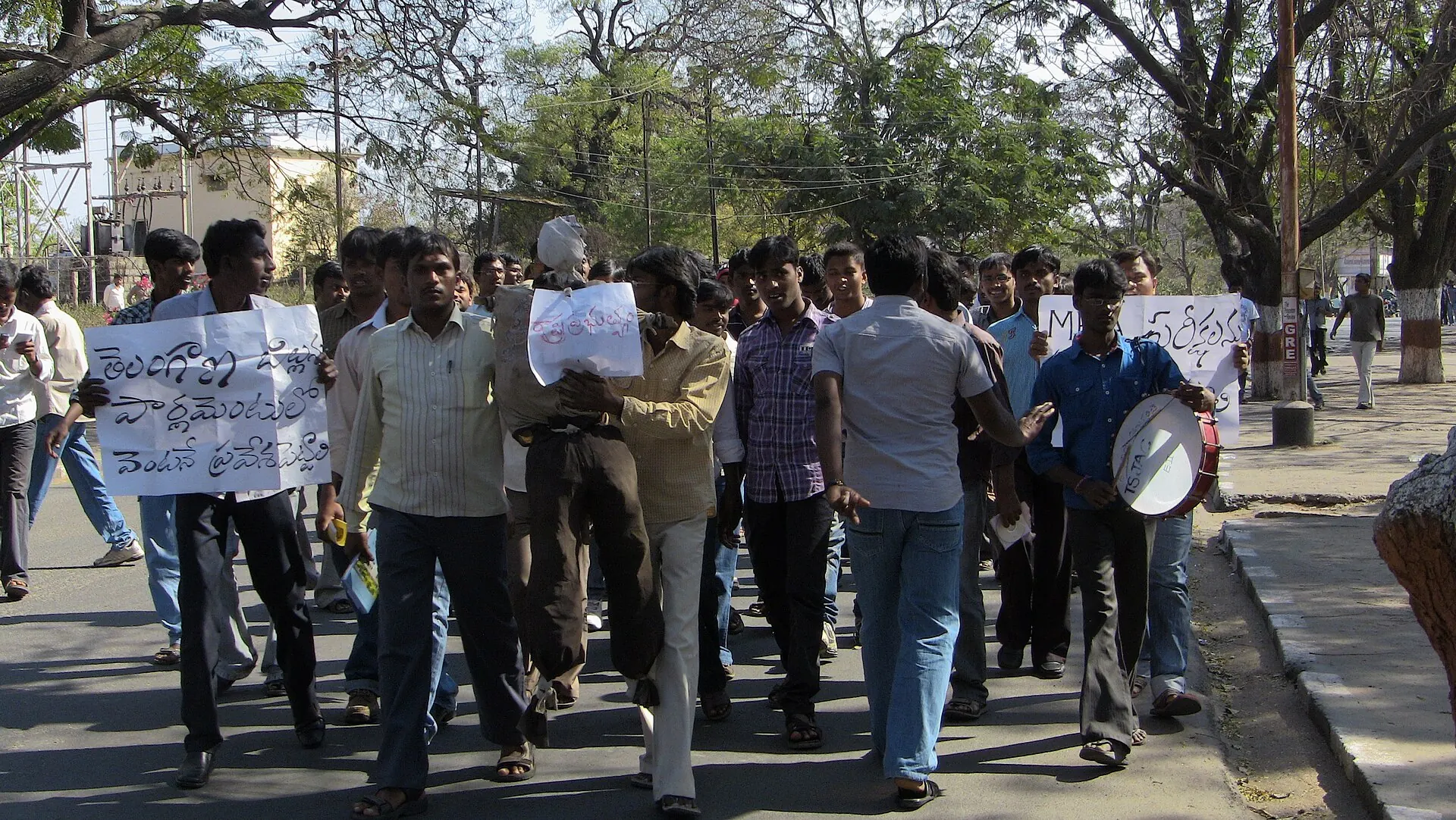 A colour photograph of a crowd of young men walking down a tree-lined road, several holding hand-written placards in Telugu and one beating a drum.