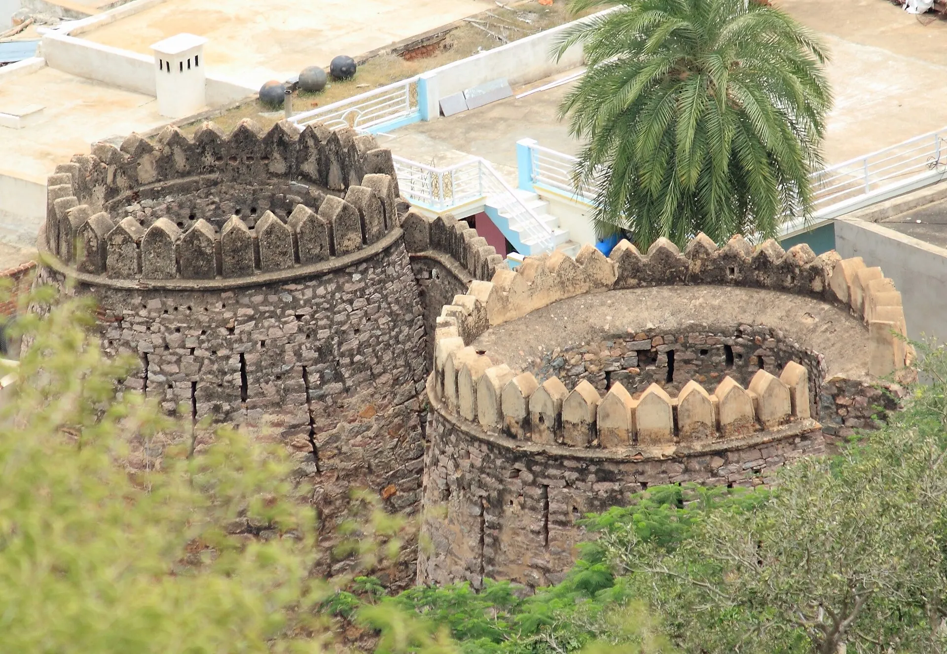 Colour photograph from above of two round stone bastions with crenellated parapets, with trees and a modern building behind.