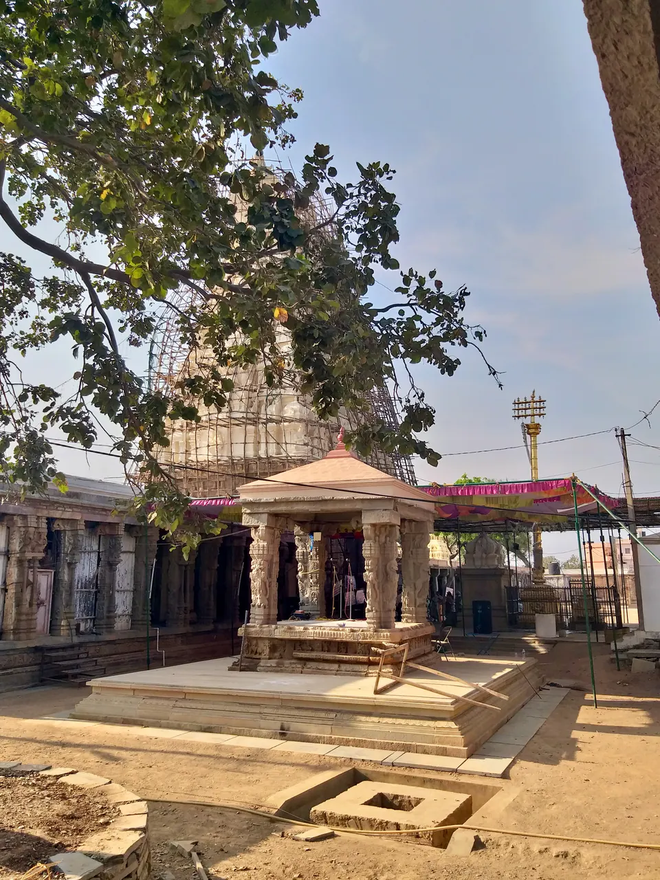 A sunny stone-paved temple courtyard with a small pillared stone pavilion on a raised base, a tower covered in scaffolding behind it and long pillared halls on both sides.