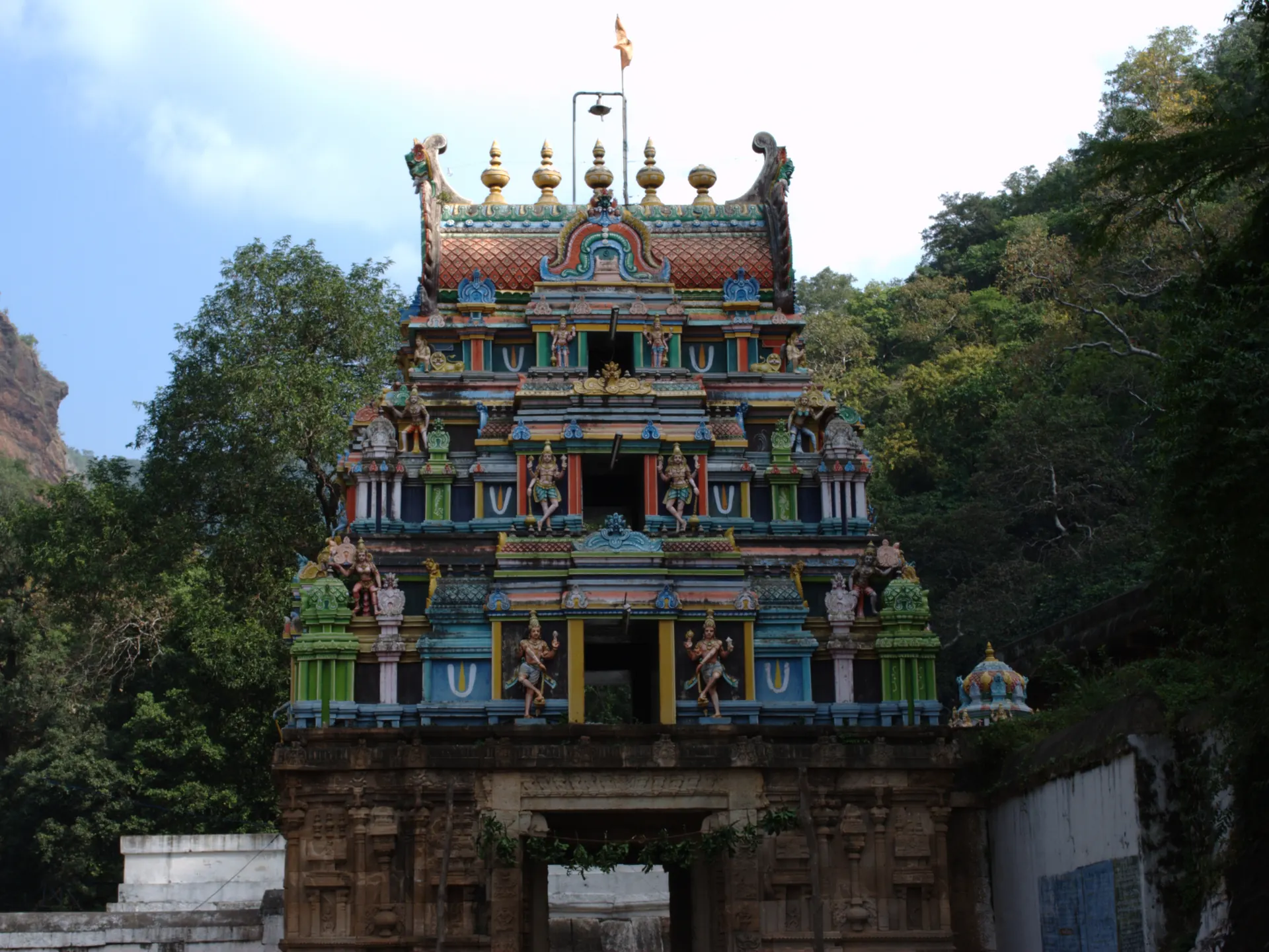 A tall painted temple gateway tower with many small sculpted figures, rising over a stone gateway, with green trees and a rocky hillside behind.