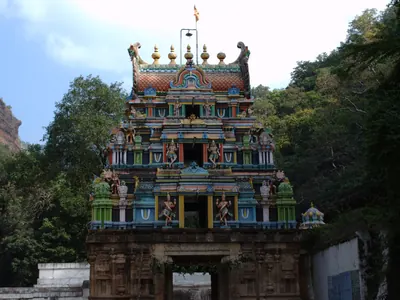 A tall painted temple gateway tower with many small sculpted figures, rising over a stone gateway, with green trees and a rocky hillside behind.