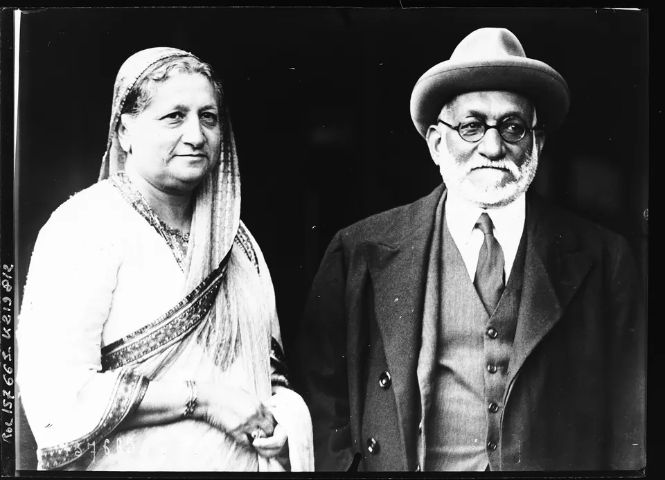 A black-and-white photograph of an elderly man with a white beard, glasses, a felt hat and a dark suit, beside an elderly woman in a light sari drawn over her head.