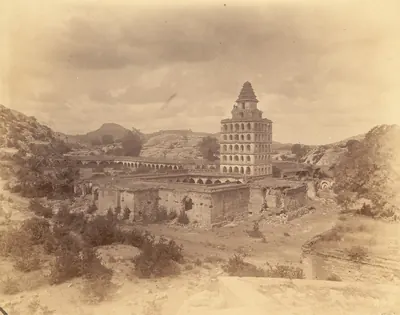An old sepia photograph of a tall square tower with many rows of small arches and a pyramid roof, standing over a ruined courtyard, with rocky hills behind.