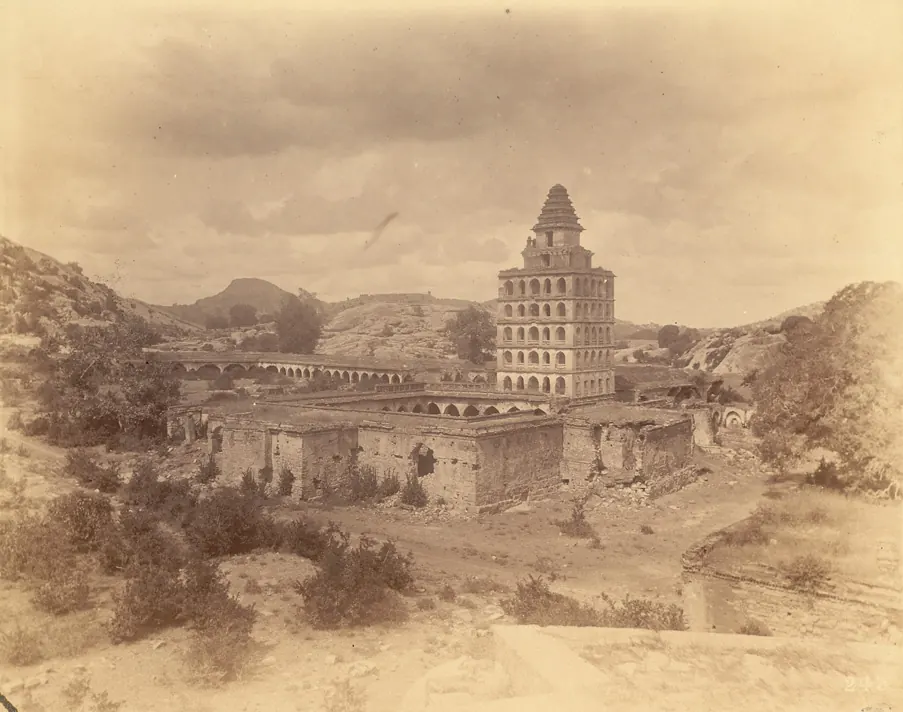 An old sepia photograph of a tall square tower with many rows of small arches and a pyramid roof, standing over a ruined courtyard, with rocky hills behind.