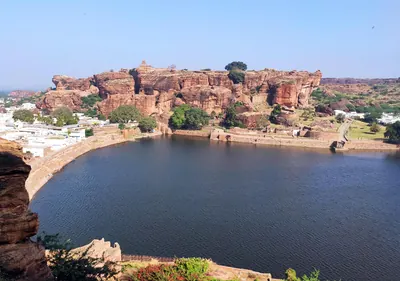 A wide blue lake curving below red sandstone cliffs topped with old walls, with white houses of the town along its shore under a clear sky.