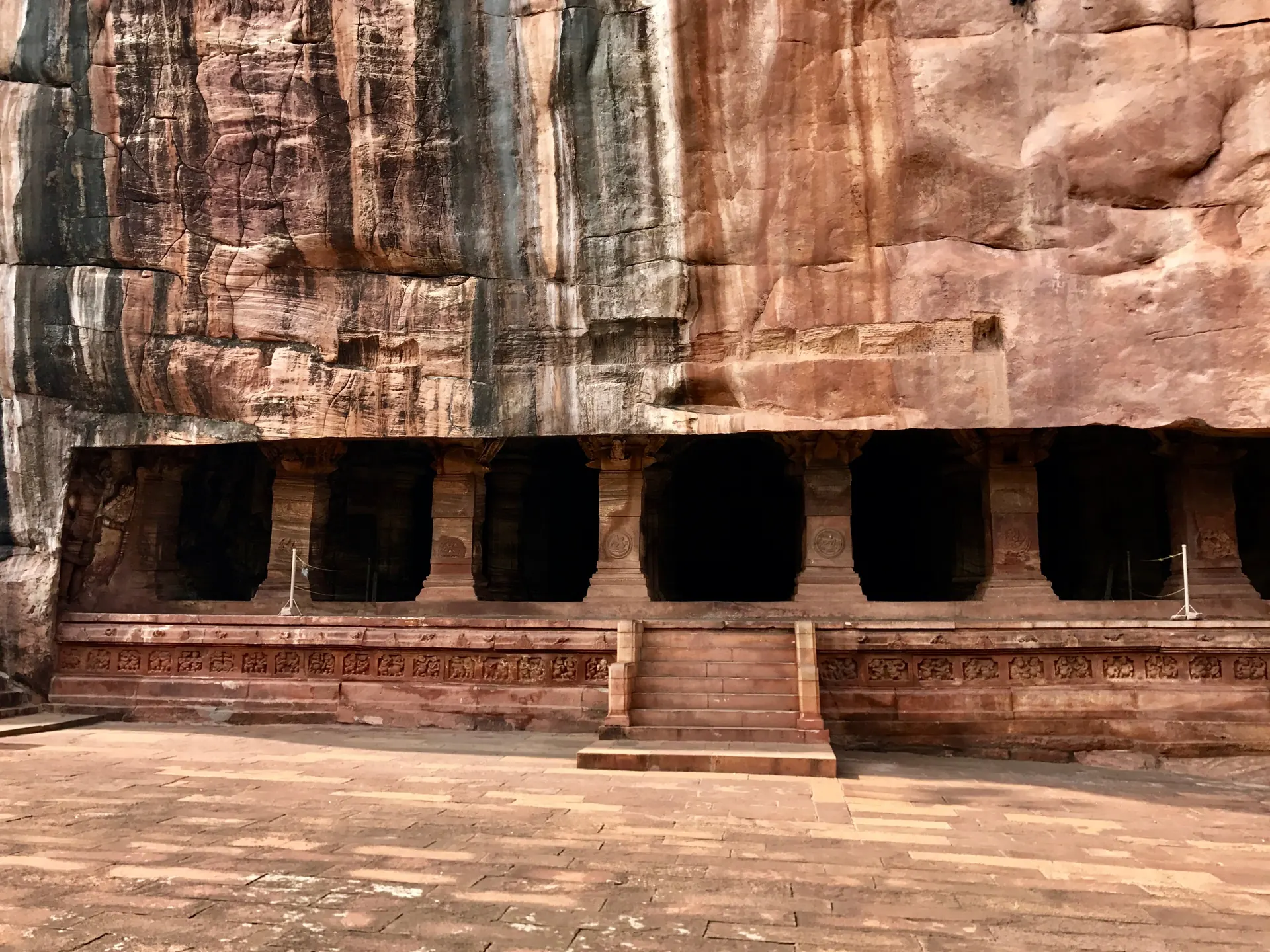 A long row of square stone pillars forming the open front of a cave cut into a tall red sandstone cliff, with steps and a carved base below.