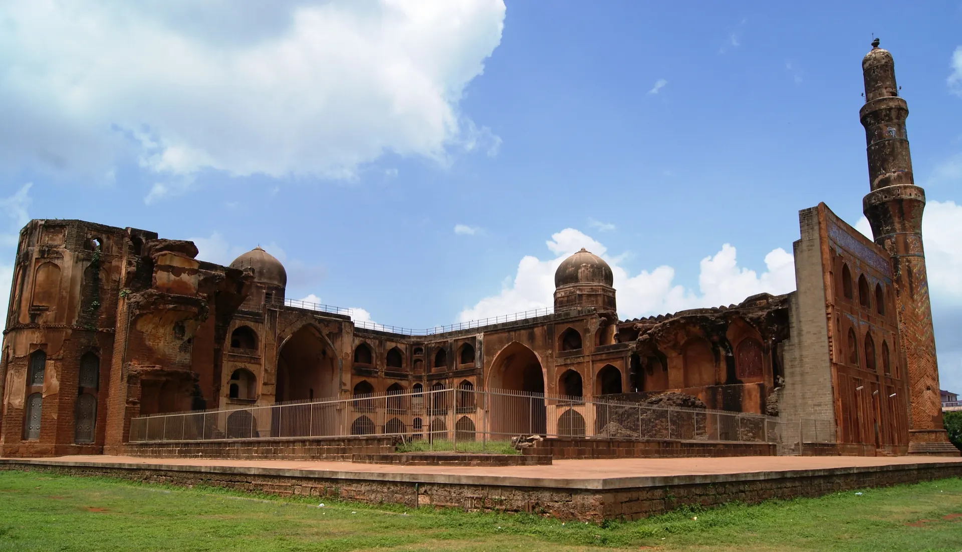 A long ruined three-storeyed building of red stone with arched windows and a tall round minaret at one end, under a blue sky with clouds.