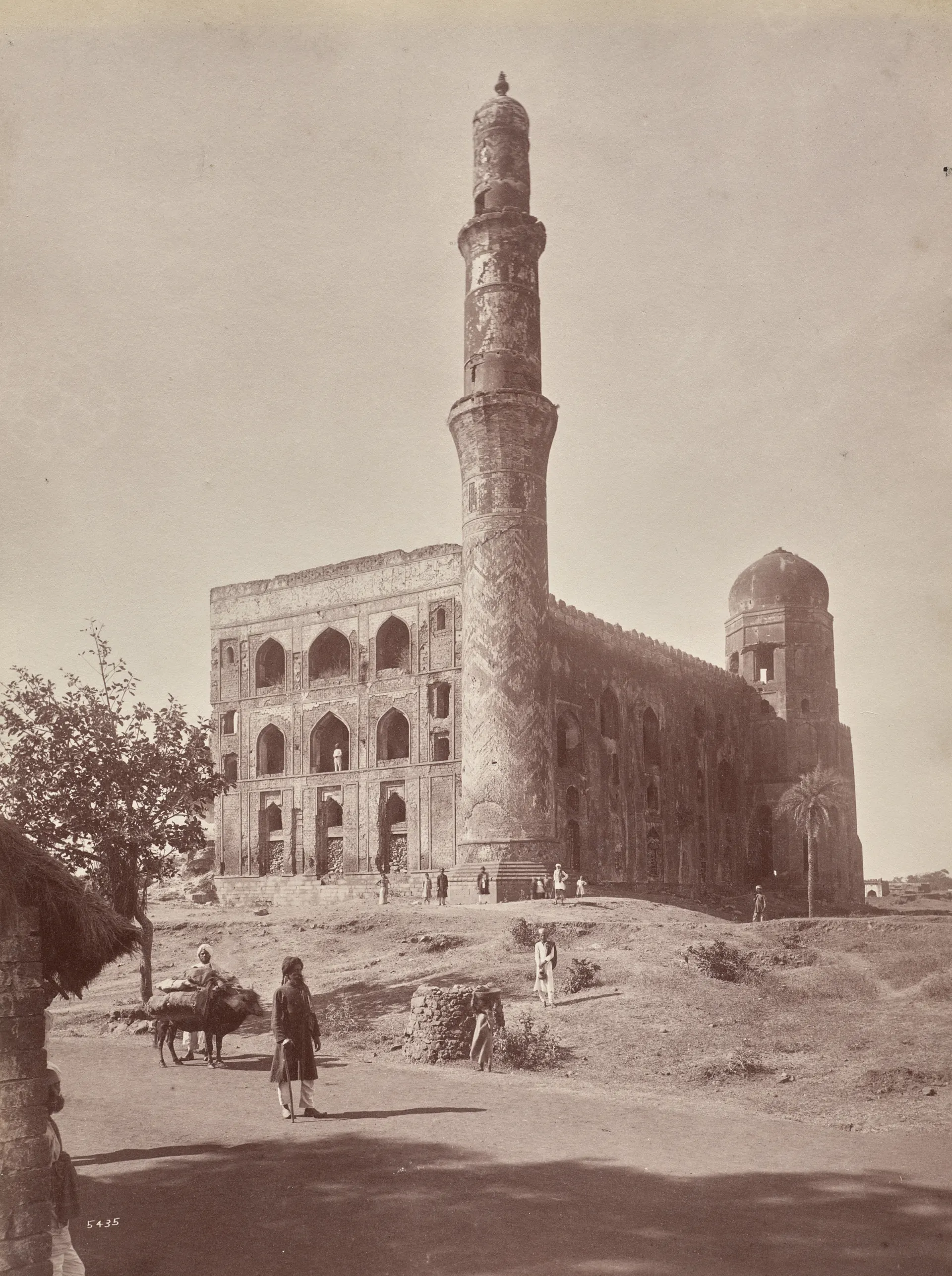 An old sepia photograph of a ruined building with a tall decorated minaret and a broken wall, with a few people standing in front.