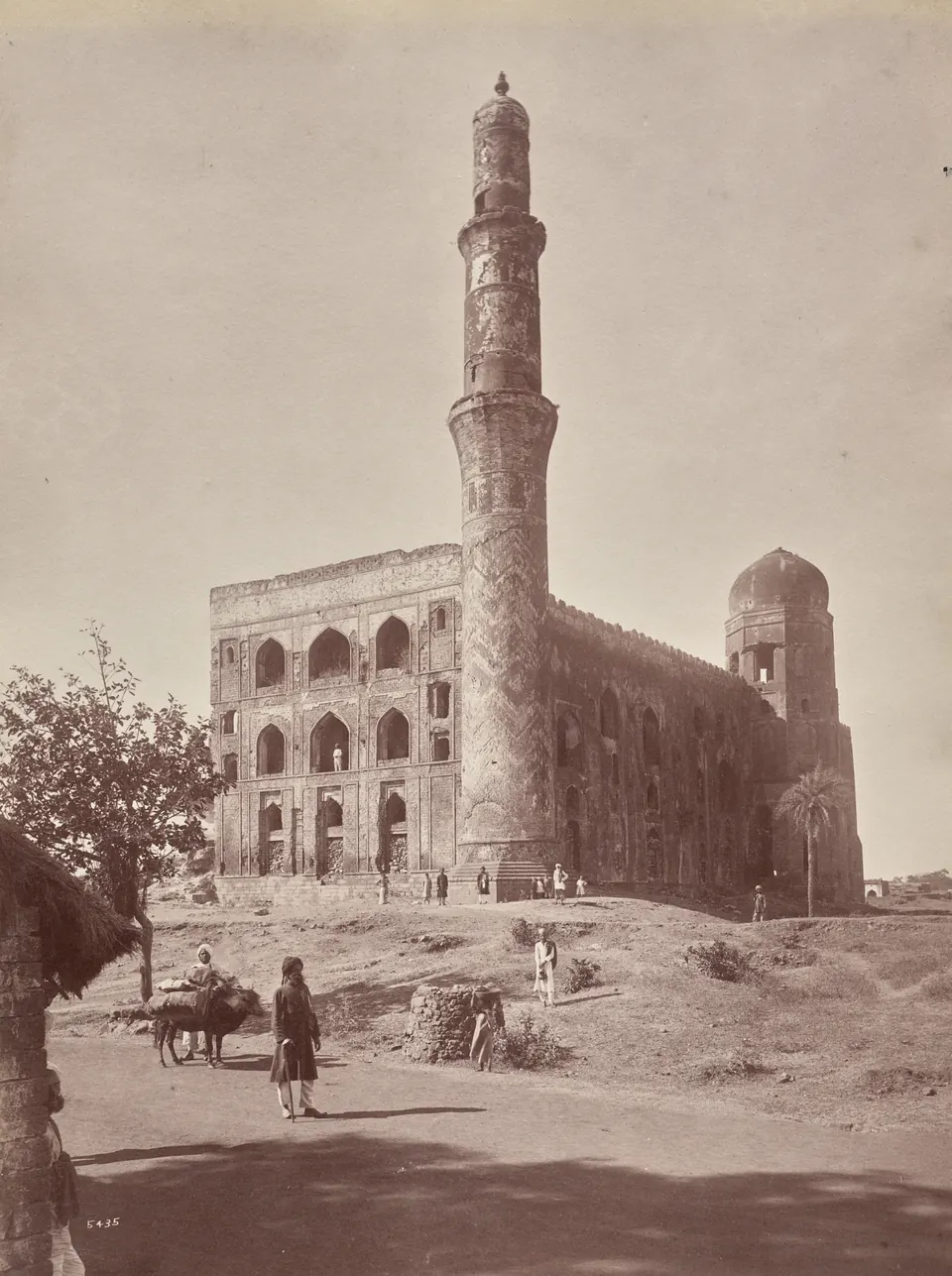 An old sepia photograph of a ruined building with a tall decorated minaret and a broken wall, with a few people standing in front.