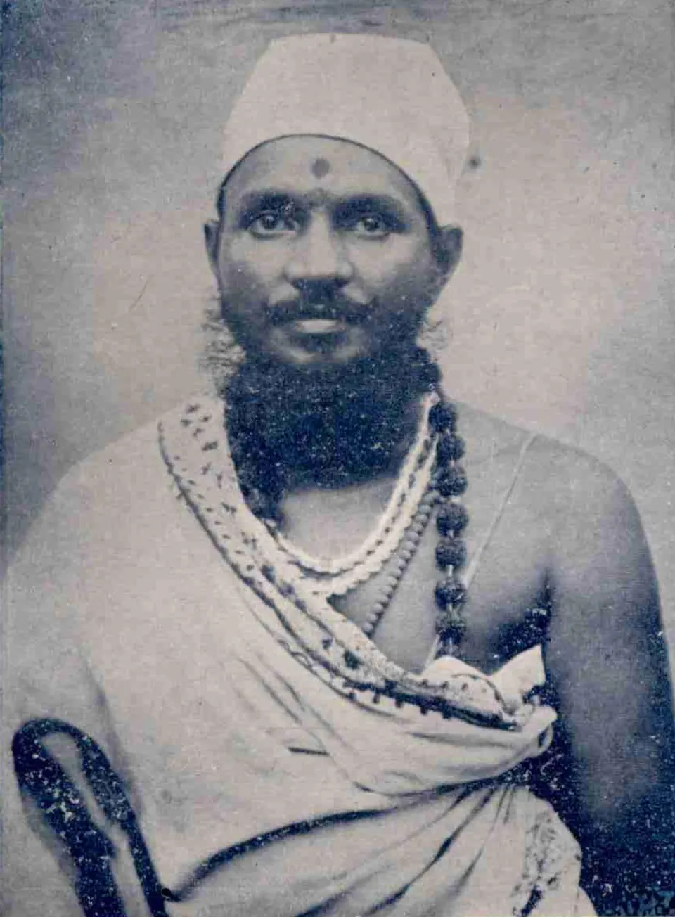 An old photograph of a bearded man in a white cap with a mark on his forehead, wearing strings of beads over a white cloth across one shoulder.