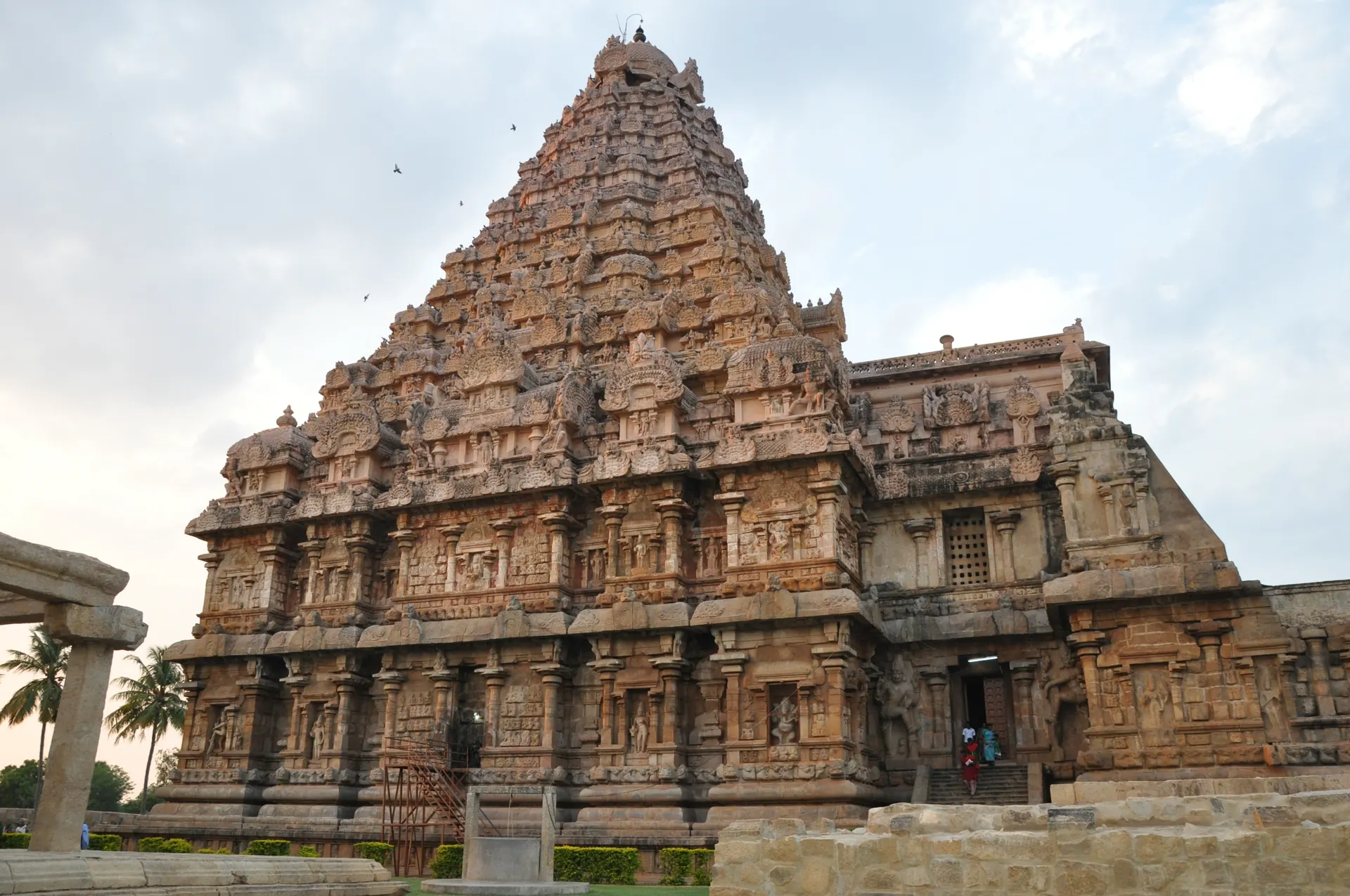 A large sandstone temple with a tall pyramid-shaped tower covered in carvings, rising behind a long pillared hall on a green lawn.