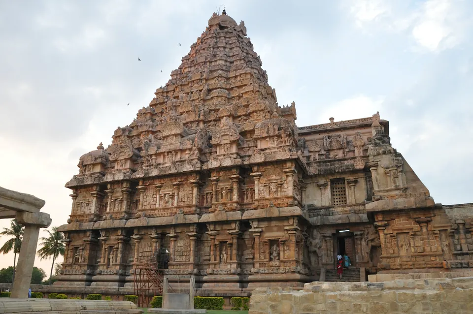 A large sandstone temple with a tall pyramid-shaped tower covered in carvings, rising behind a long pillared hall on a green lawn.