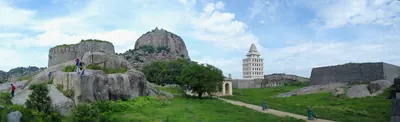 A wide view of a green fort enclosure with big boulders and stone walls; a rocky hill with a fort on top rises behind, and a white many-arched tower stands on the right.
