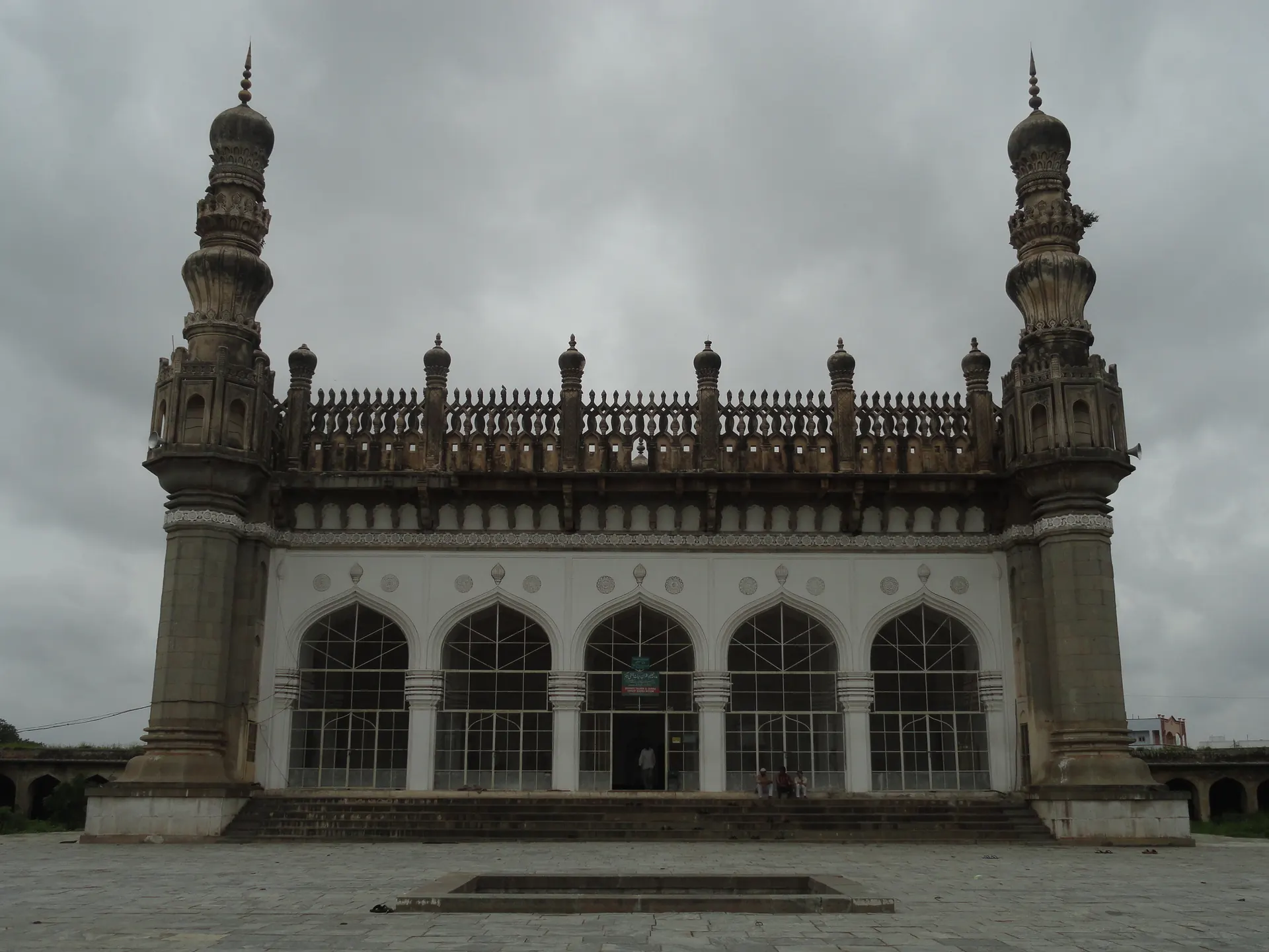 A long grey stone mosque with five arched openings, a carved parapet along the roof and a tall slender minaret at each end, under a cloudy sky.