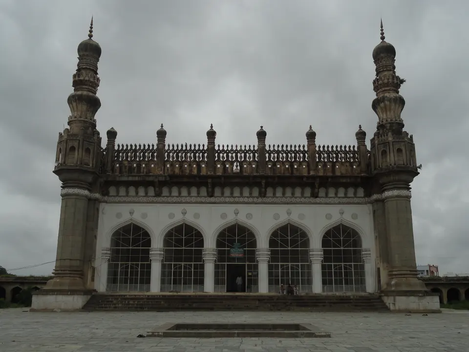 A long grey stone mosque with five arched openings, a carved parapet along the roof and a tall slender minaret at each end, under a cloudy sky.