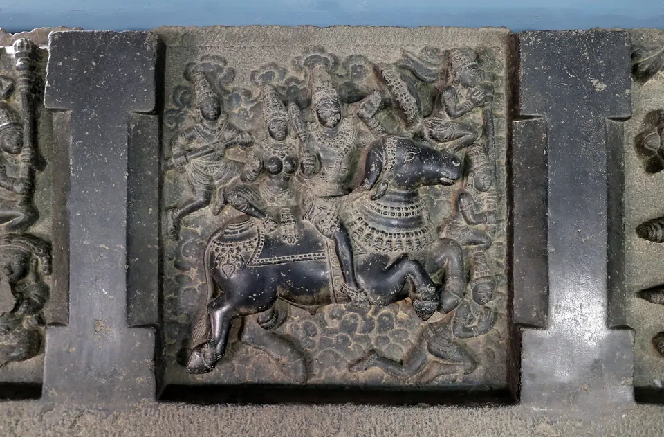 A square black stone panel framed by a raised border, carved with a figure riding an animal among smaller figures, set in a long stone beam.
