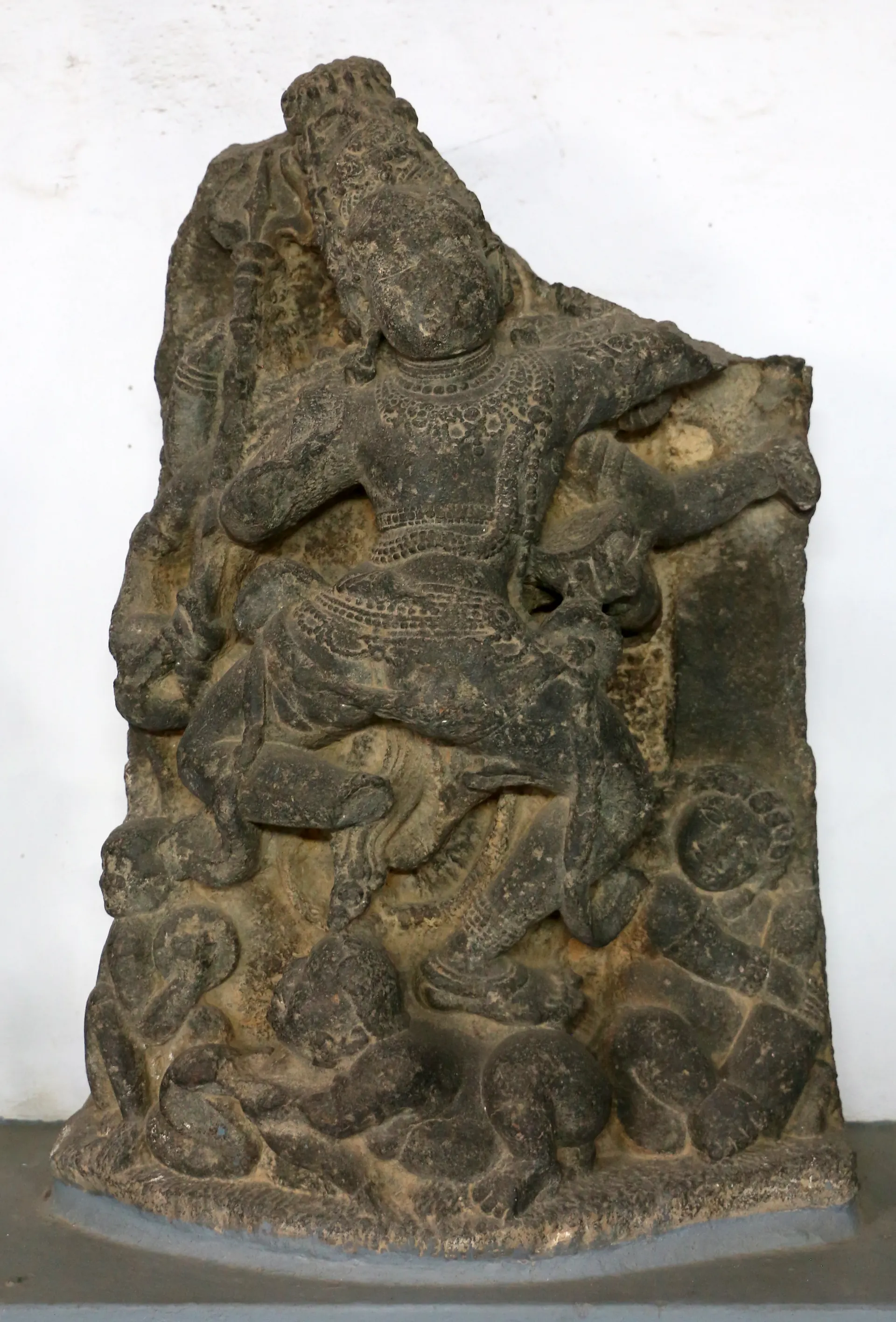 A dark grey stone relief of a many-armed dancing figure with a tall crown, worn and broken at the top, standing on a museum plinth.