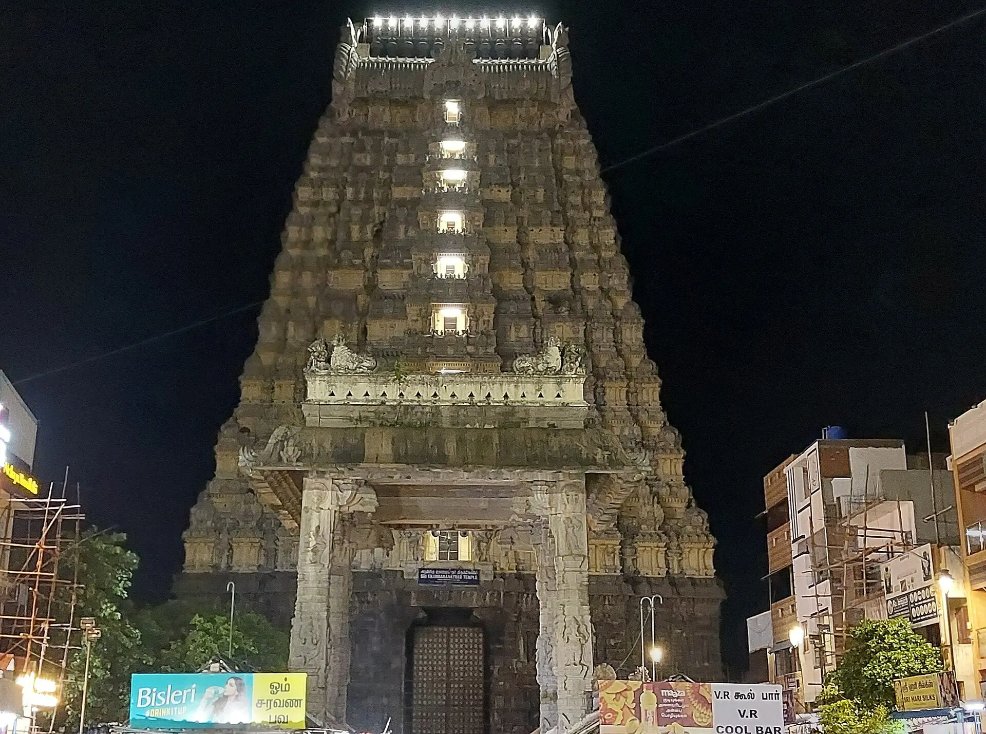 A tall stepped stone gateway tower lit by lamps against a black night sky, with a carved entrance at its foot and buildings beside it.