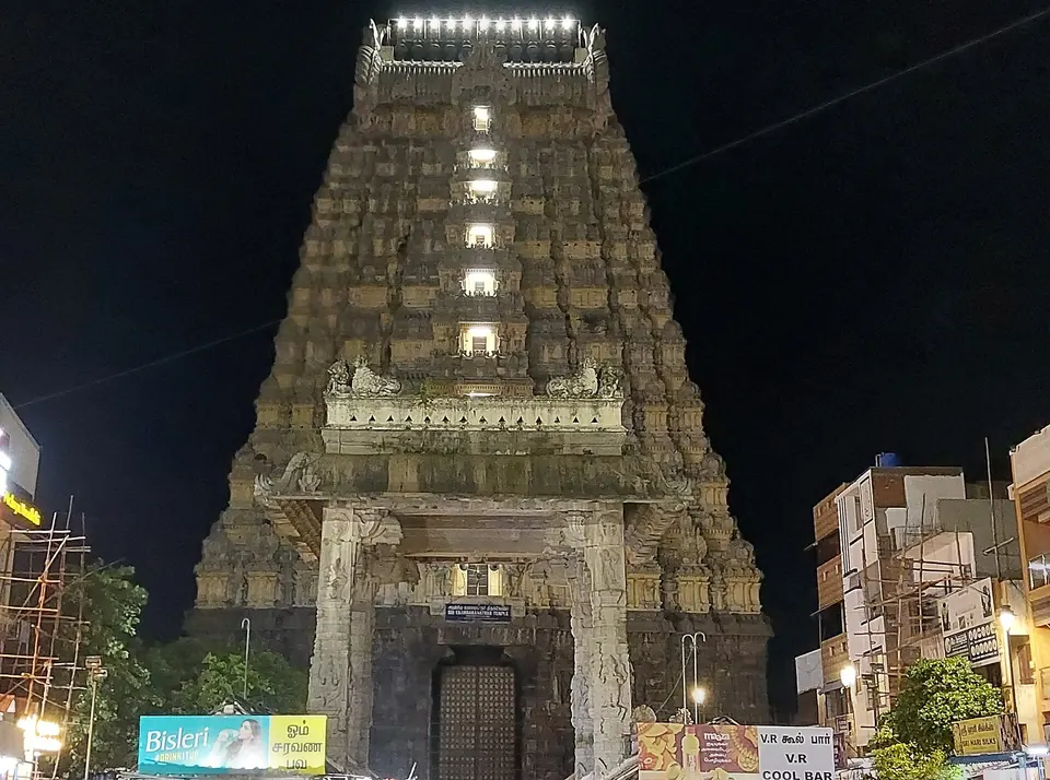 A tall stepped stone gateway tower lit by lamps against a black night sky, with a carved entrance at its foot and buildings beside it.