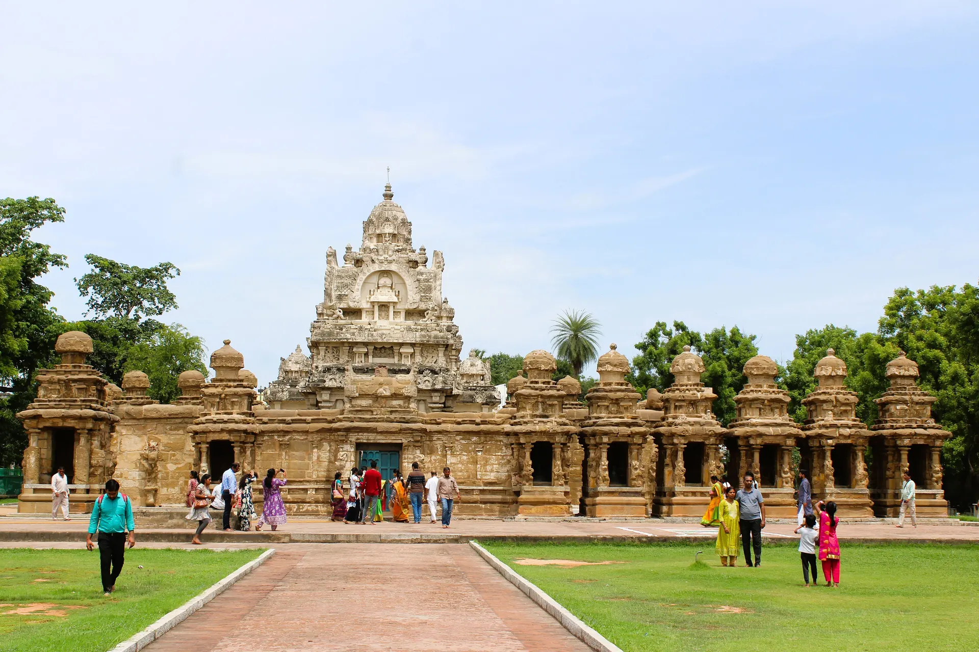 A sandstone temple with a stepped pyramid tower behind a long low wall lined with small shrines, approached by a red path across green lawns, with visitors in front.