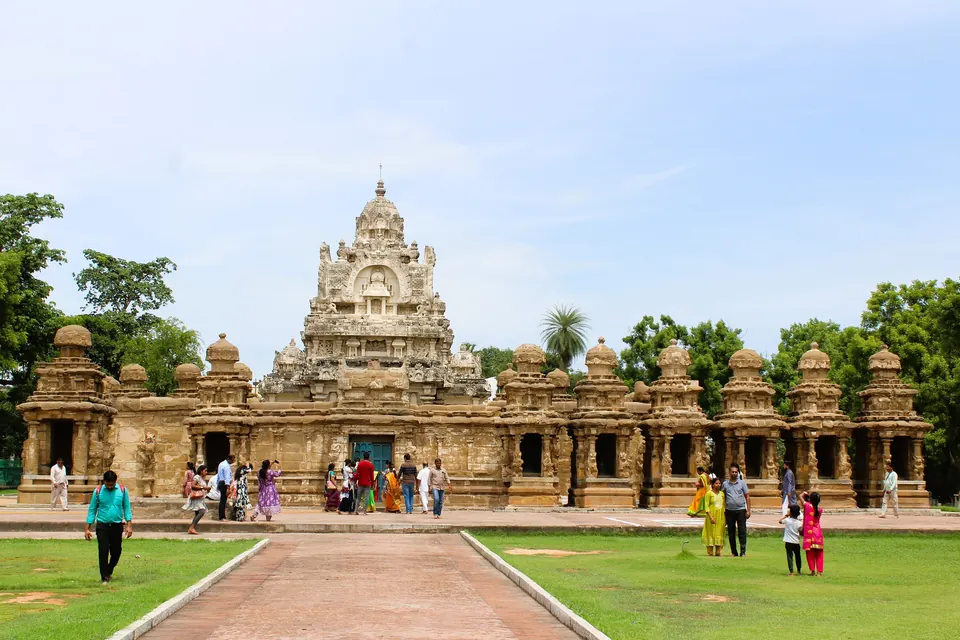 A sandstone temple with a stepped pyramid tower behind a long low wall lined with small shrines, approached by a red path across green lawns, with visitors in front.