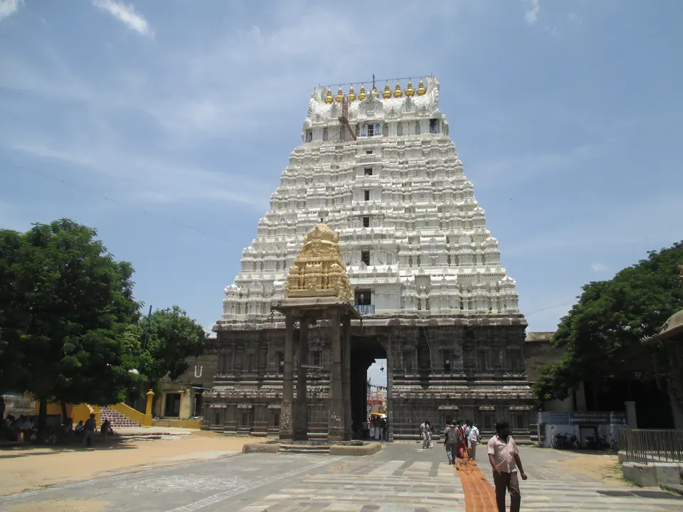 A tall white temple gateway tower with many tiers of sculpture above a dark stone base, with a smaller golden-roofed shrine and a few people in the paved court in front.