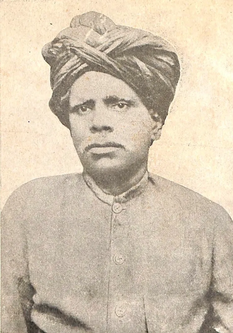 An old sepia photograph of a man with a large turban and a stern face, wearing a plain buttoned coat.