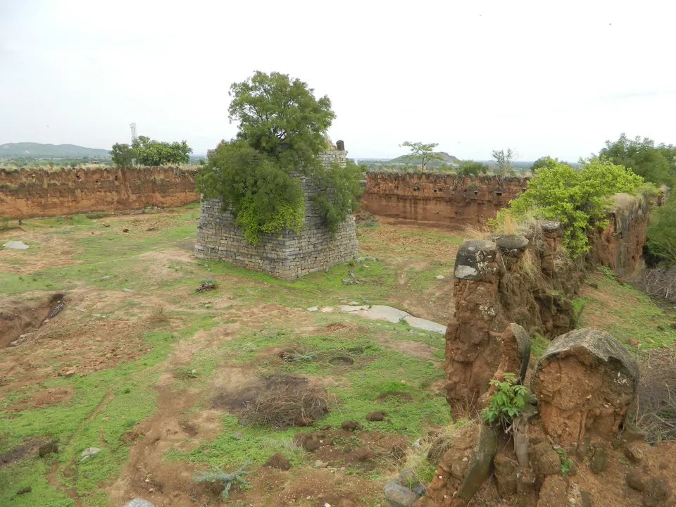 A grassy enclosure inside long walls of dark stone, with a ruined square bastion overgrown by a tree and broken rock in front.
