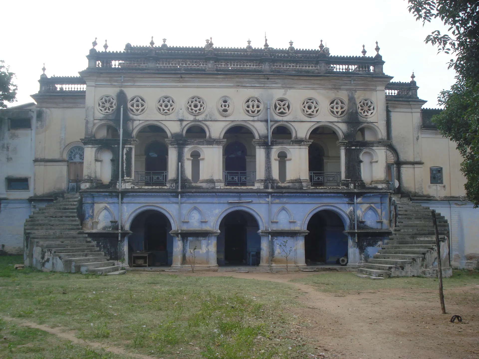 A large two-storeyed palace painted blue and white, with three arched doorways, rows of round windows above, and staircases at both ends, behind an open dusty yard.
