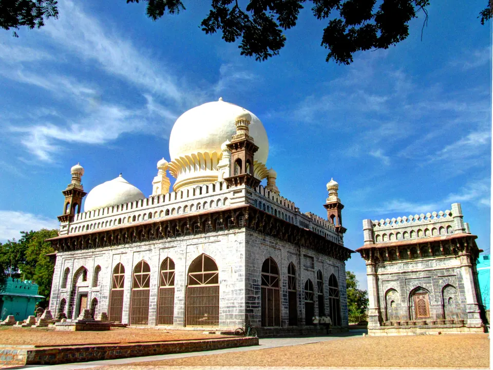 A large square stone tomb with rows of arches, small corner towers and a big white onion dome, with a smaller arched building beside it under a blue sky.
